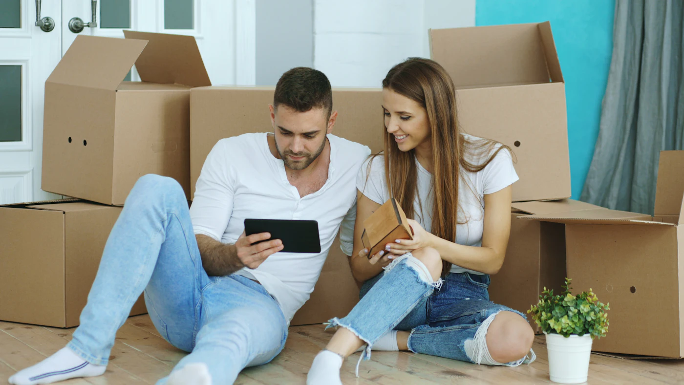 Two young people sitting on the floor of a new flat among moving boxes, looking at a tablet together
