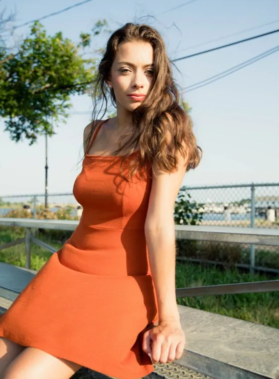 A young woman in an orange dress sitting on a sunny outdoor bench, looking at the camera