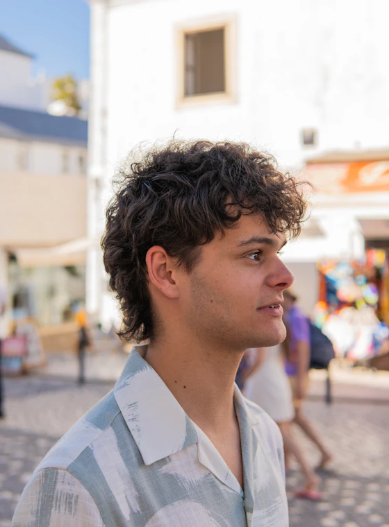 A young man with curly hair standing in a sunny old-town street