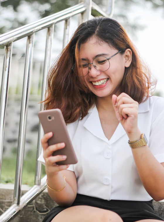 A young woman in glasses smiling at her phone outdoors